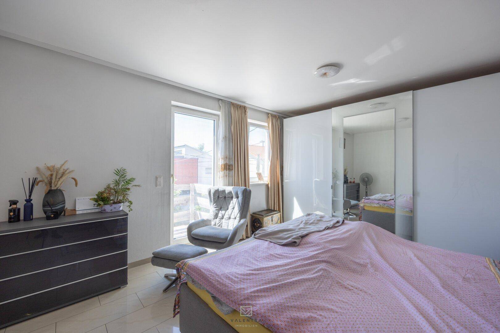 Helles Schlafzimmer mit Bett, Kommode, Sessel und Spiegelschrank, mit Zugang zu einem Fenster.