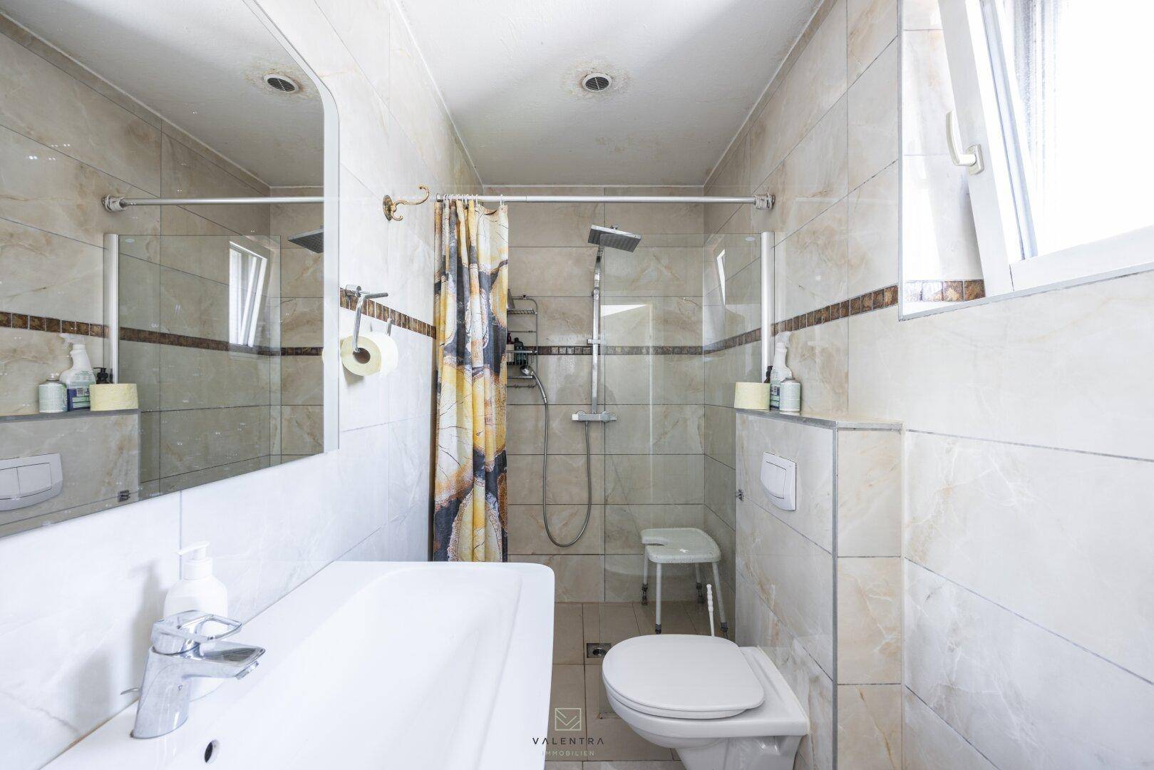 Modernes Badezimmer mit Dusche, Toilette, Waschbecken und Fenster, komplett gefliest in hellen Tönen.