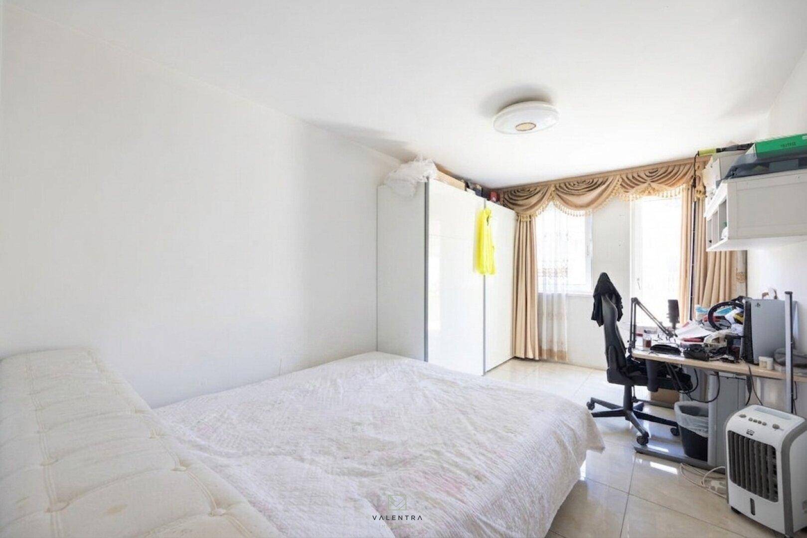 Helles Schlafzimmer mit Bett, Kleiderschrank, Schreibtisch und Fenster mit Vorhängen.
