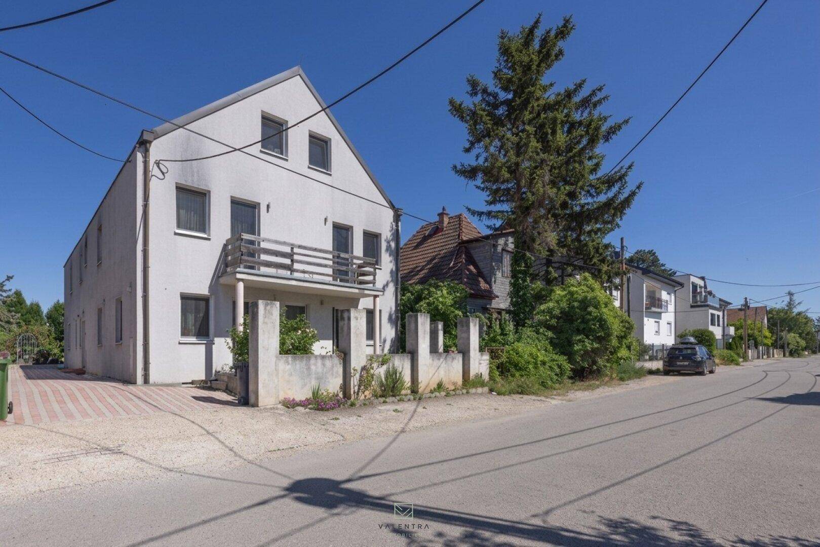 Ansicht des Hauses von der Straße mit Zufahrt und umliegender Bebauung unter blauem Himmel.