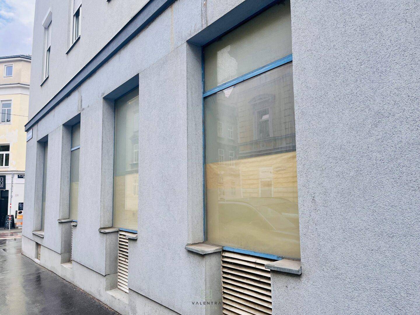 Gebäudefassade mit großen, abgedeckten Fenstern und Lüftungsschlitzen im unteren Bereich entlang der Straße.