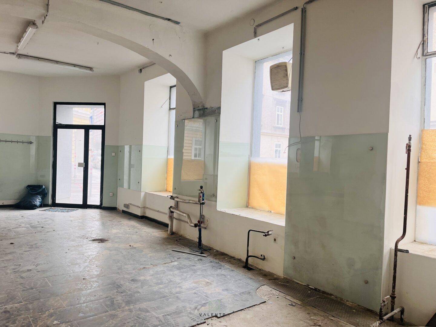 Heller Innenraum mit großen Fenstern, sichtbaren Rohren und einem Eingangsbereich, der zur Straße führt.