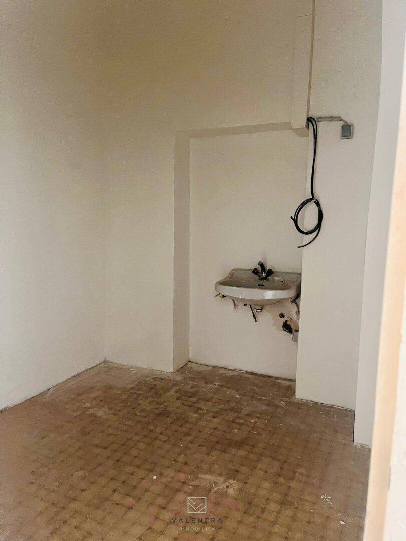 Kleiner Raum mit einem Waschbecken und sichtbaren Leitungen, der als Toilette oder Abstellraum dienen könnte.