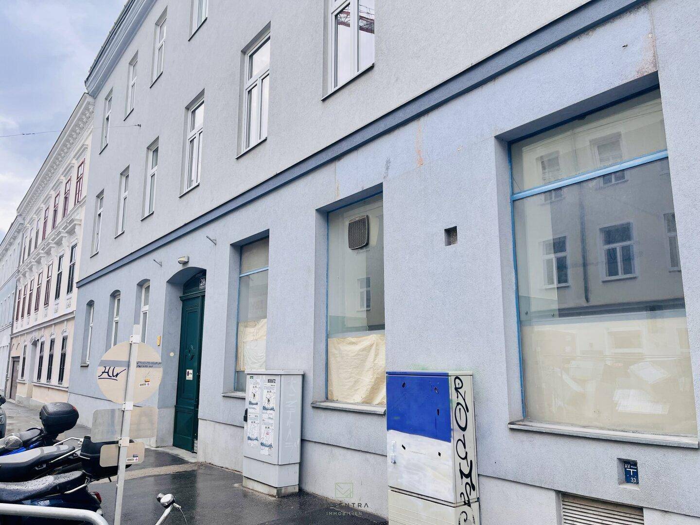 Fassade eines älteren Gebäudes mit mehreren Fenstern und einem grünen Eingang an einer belebten Straße.