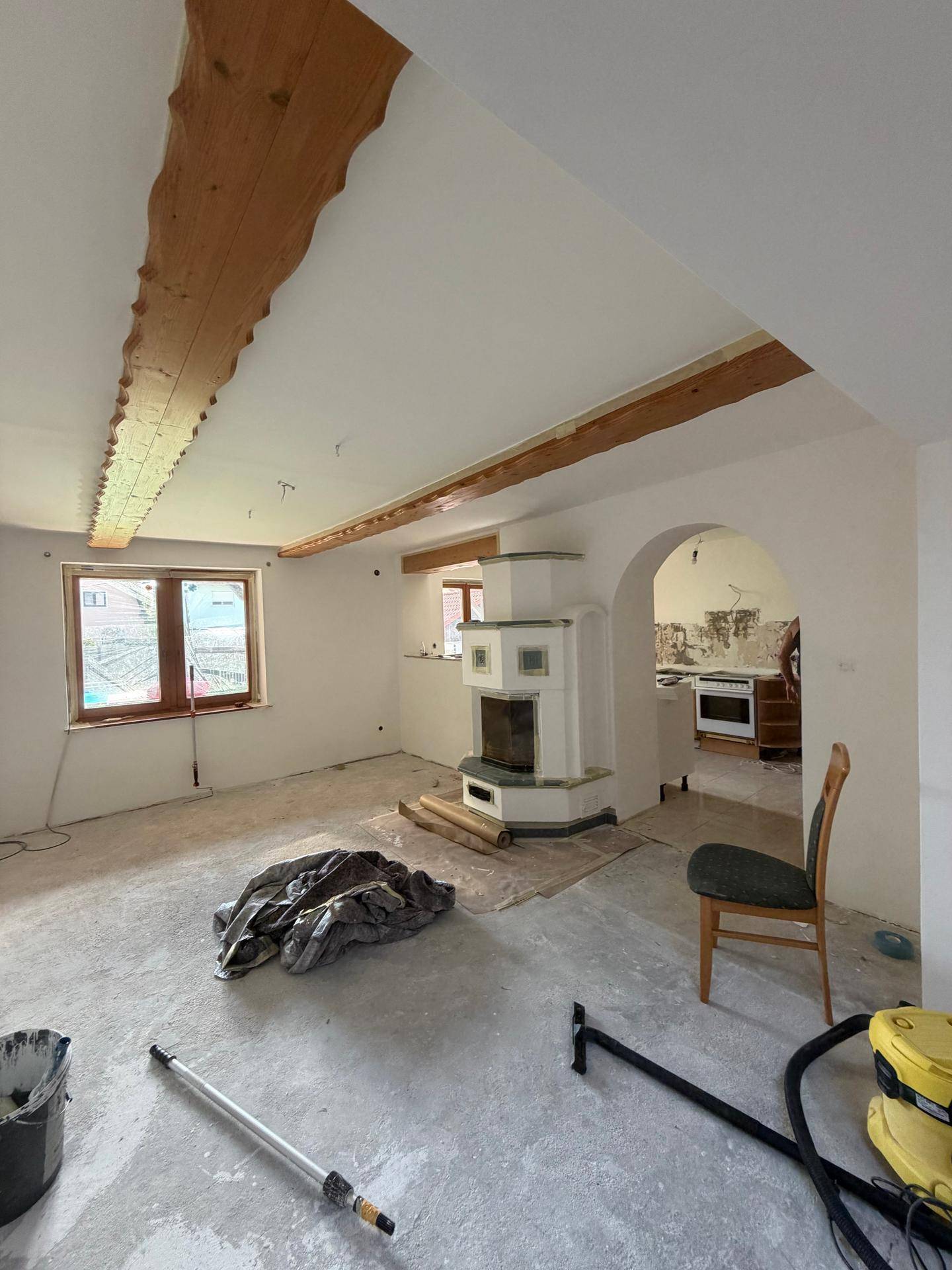 Geräumiges Wohnzimmer im Umbau mit Kachelofen, Holzbalken und offenem Zugang zur Küche.