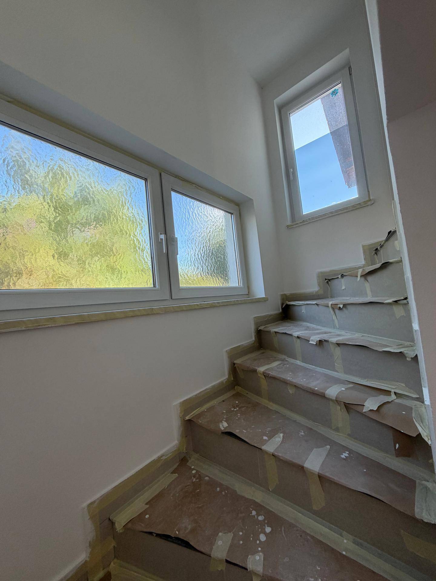 Helles Treppenhaus im Umbau mit mehreren Fenstern und Blick ins Grüne, viel Tageslicht.