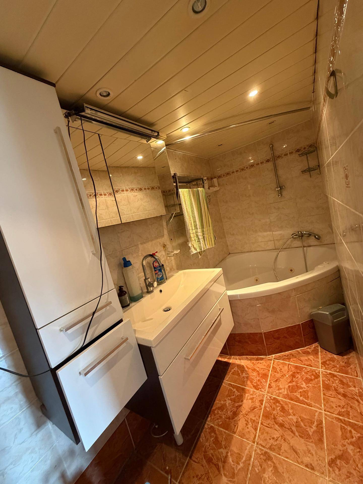 Badezimmer mit Eckbadewanne, Waschtisch und hellbraunen Fliesen, funktional eingerichtet.