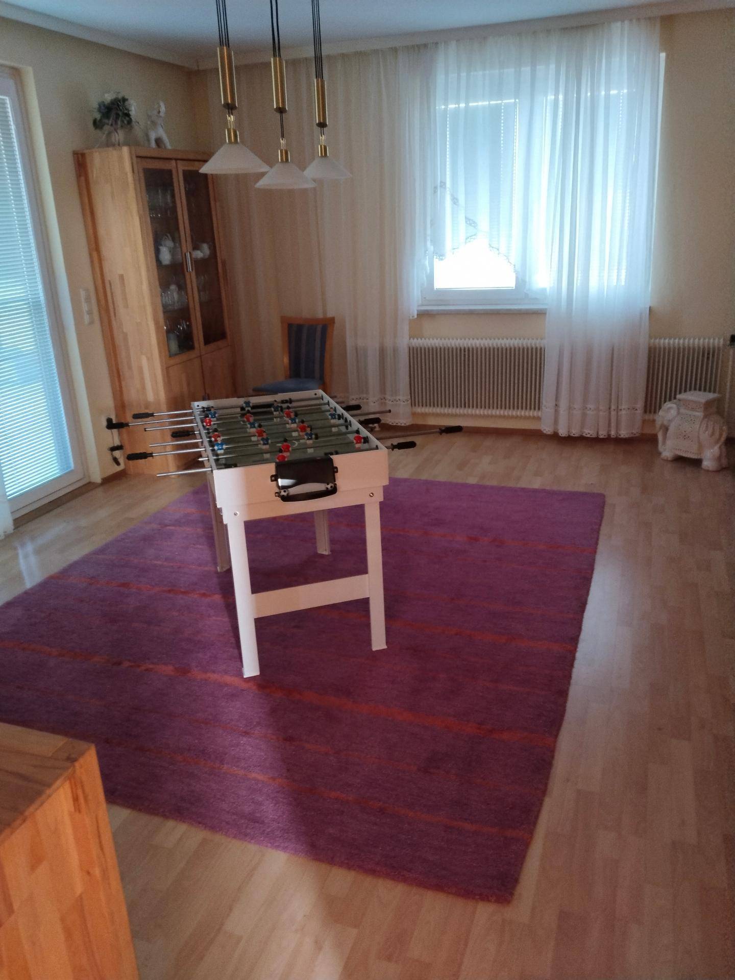 Helles Wohnzimmer mit Holzboden, großem Fenster und einem Tischfußballspiel in der Mitte des Raumes.