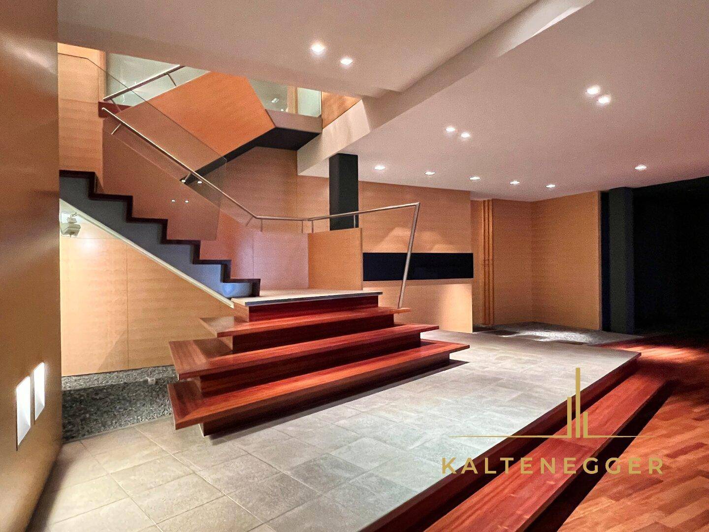Elegante Eingangshalle mit breiter Holztreppe, Glasgeländer und verschiedenen Bodenbelägen unter hoher Decke.