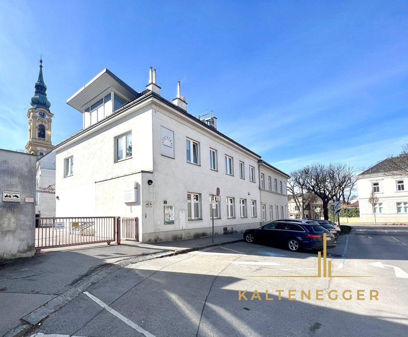 Gebäudeansicht von der Straße mit Parkplätzen und einem historischen Kirchturm im Hintergrund unter blauem Himmel.