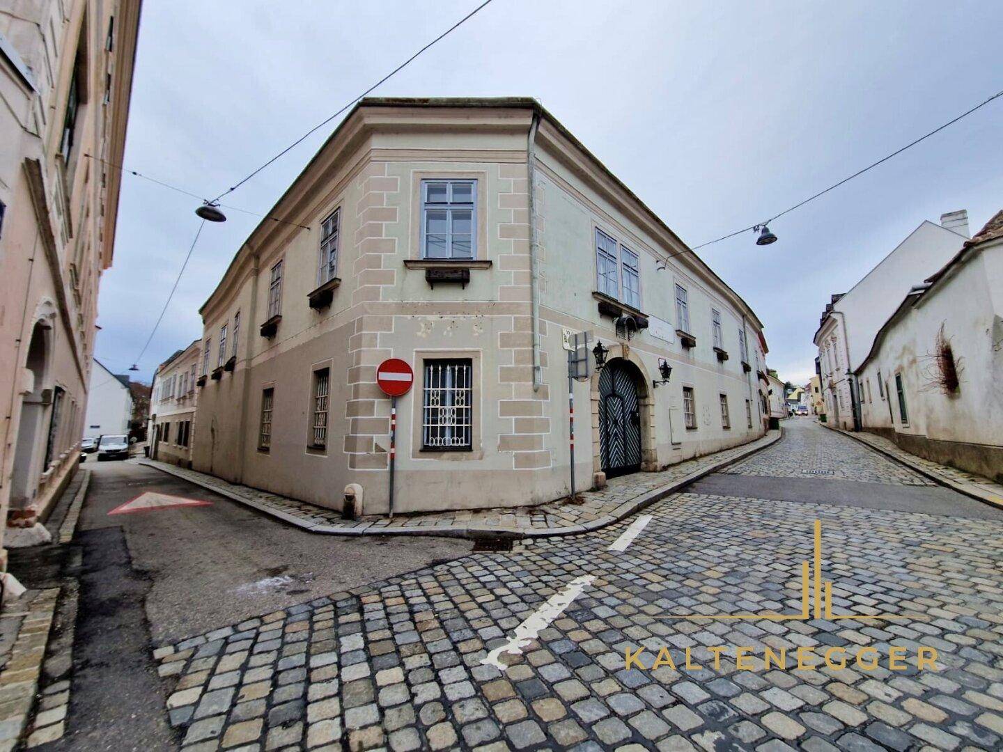 Weitwinkelansicht des historischen Eckgebäudes an einer gepflasterten Gasse unter bewölktem Himmel.