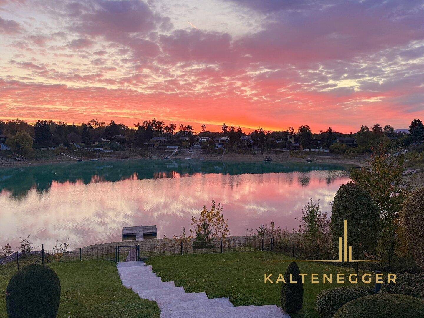 Malerischer Sonnenuntergang über dem See, eingefangen vom grünen Gartenbereich des Anwesens.