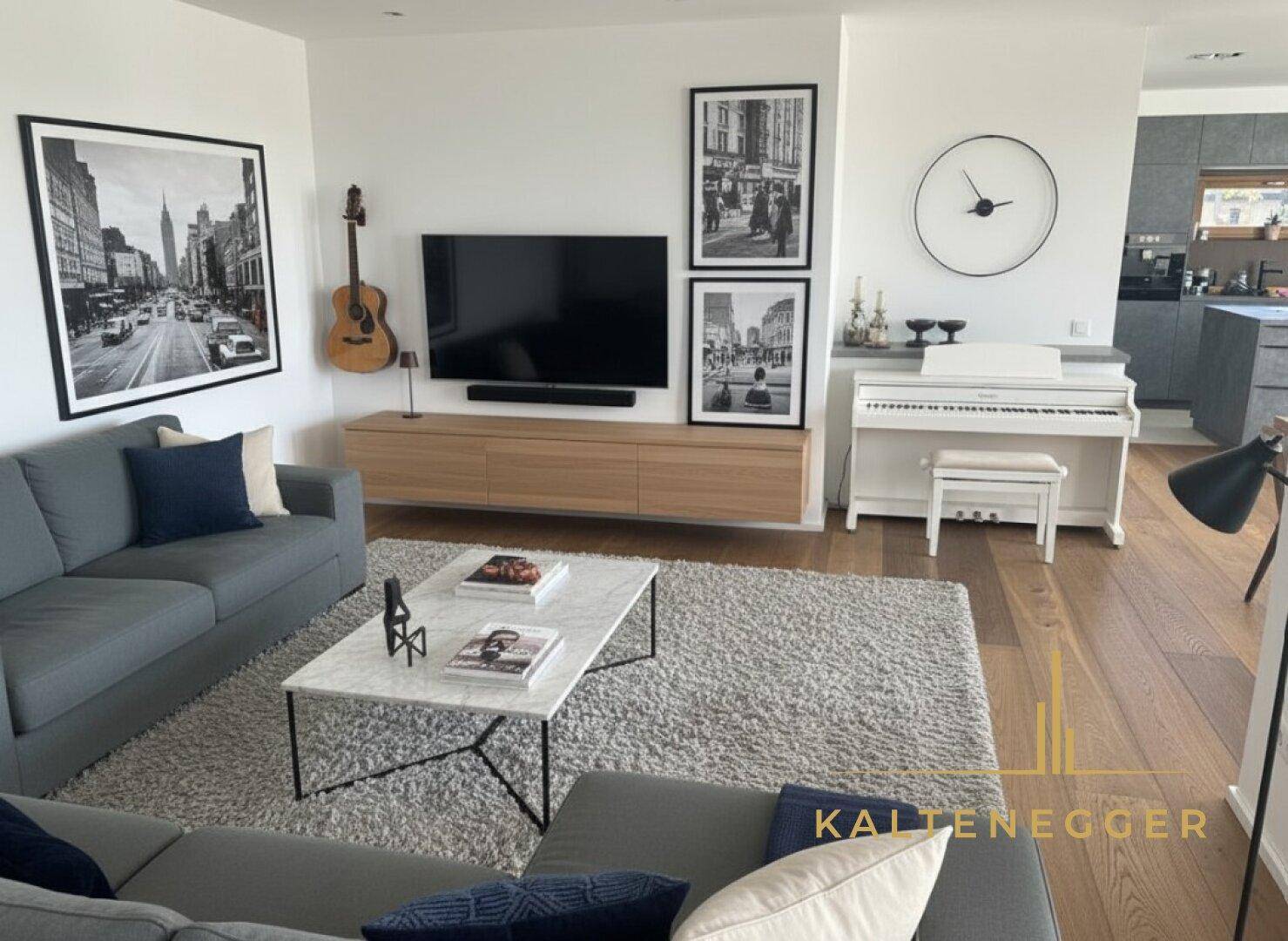 Gemütliches Wohnzimmer mit Sofa, Teppich, Klavier und Holzboden, offen zur Küche.