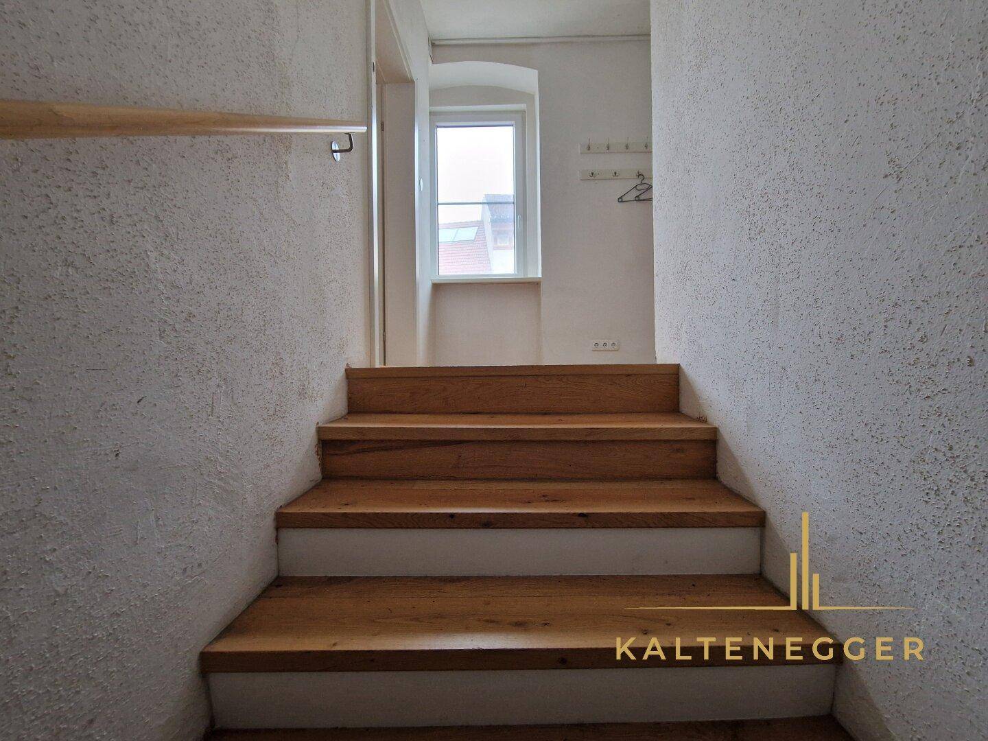 Holztreppe führt zu einem oberen Stockwerk mit einem kleinen Fenster und weiß verputzten Wänden.
