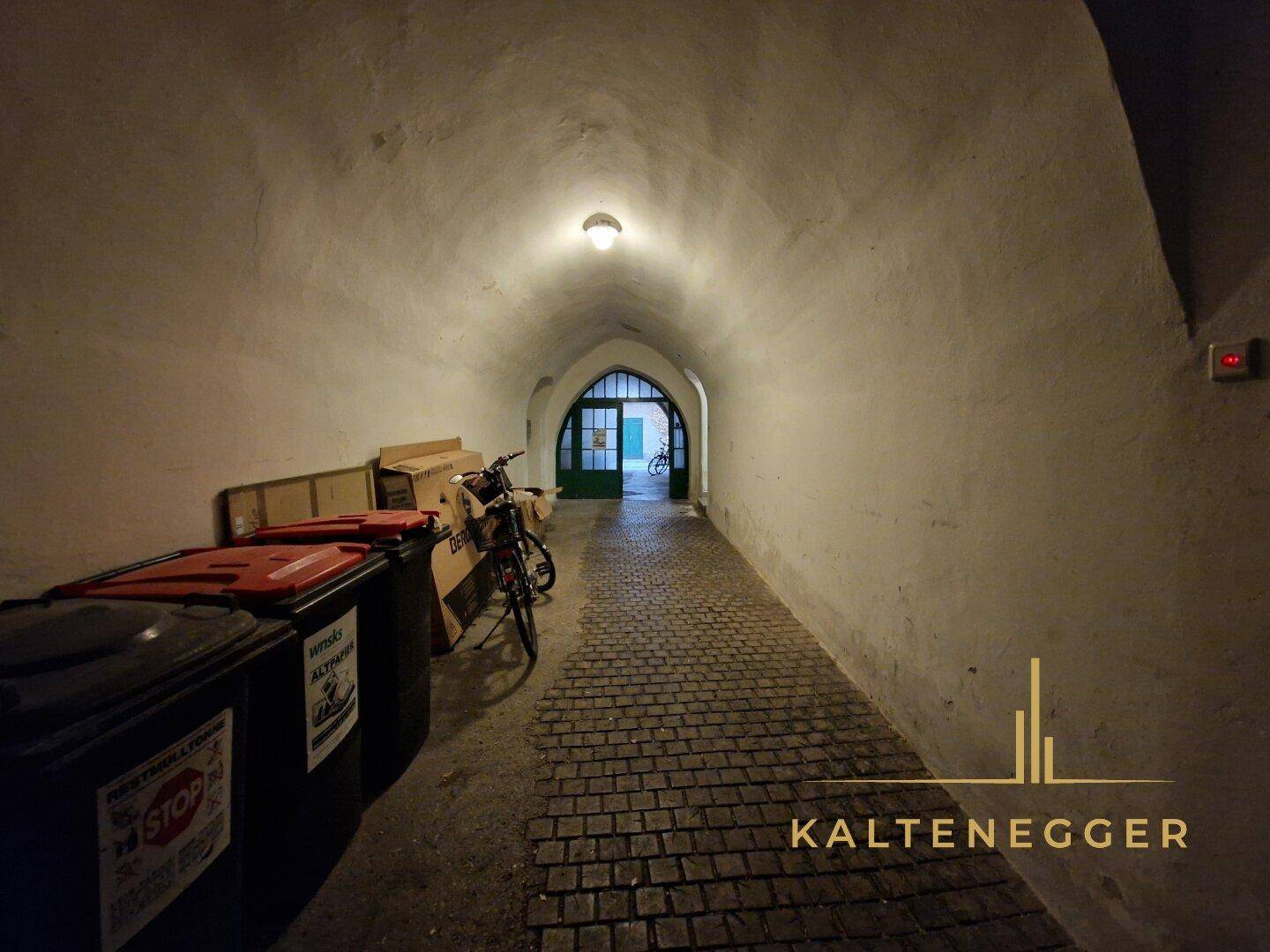 Langer, gewölbter Durchgang mit Kopfsteinpflaster und Mülltonnen, der zum Hof führt.