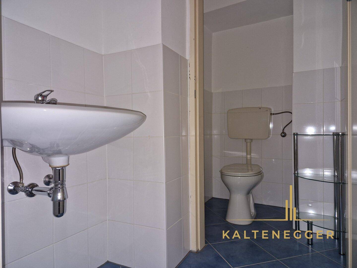 Badezimmer mit Waschbecken und Toilette, ausgestattet mit weißen Wandfliesen und blauen Bodenfliesen.