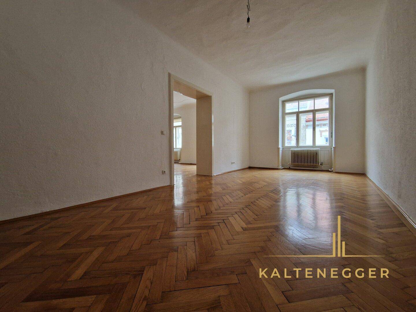Helles Zimmer mit Fischgrätparkett und einem großen Fenster, das viel Tageslicht hereinlässt.