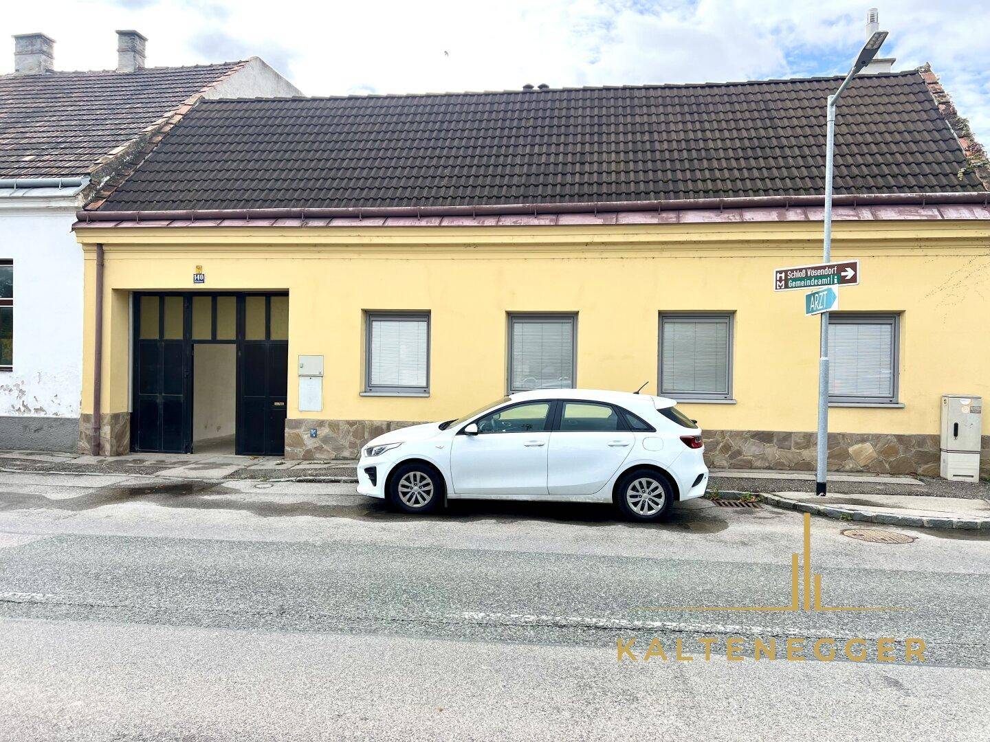 Traditionelles gelbes Haus an einer Straße mit Parkplatz und angrenzenden Gebäuden.