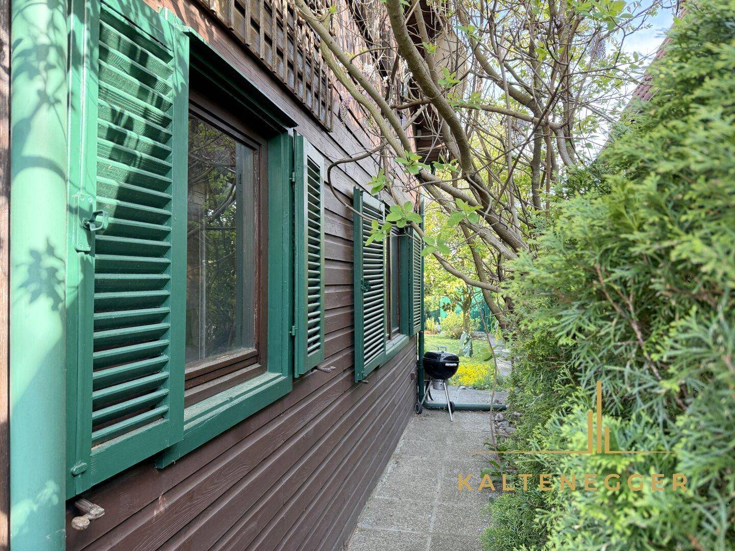 Seitenansicht des Holzhauses mit grünen Fensterläden und einem Grillbereich im Freien.