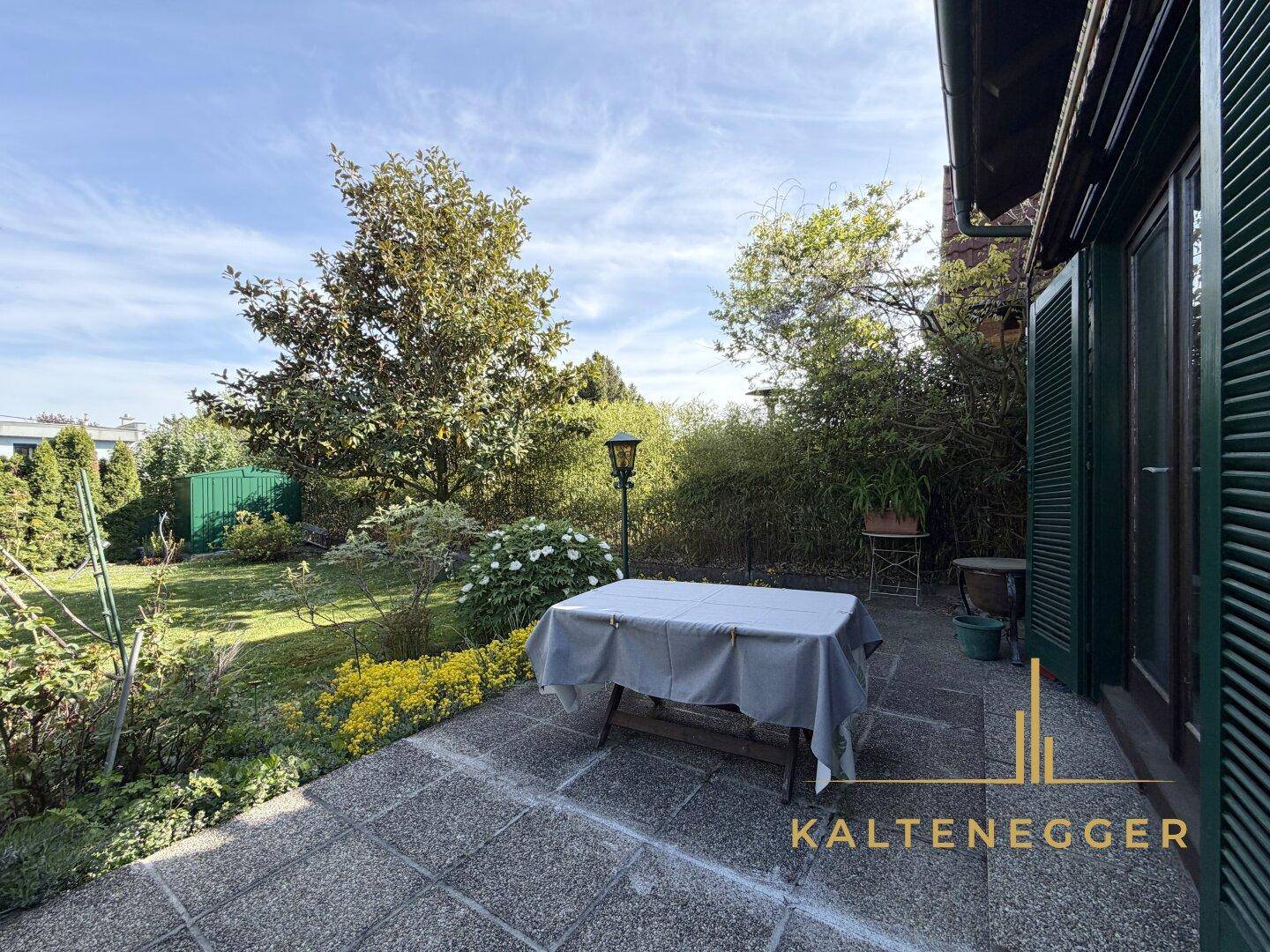 Gepflasterte Terrasse mit Tisch und Blick auf den grünen Garten und die umliegenden Bäume.