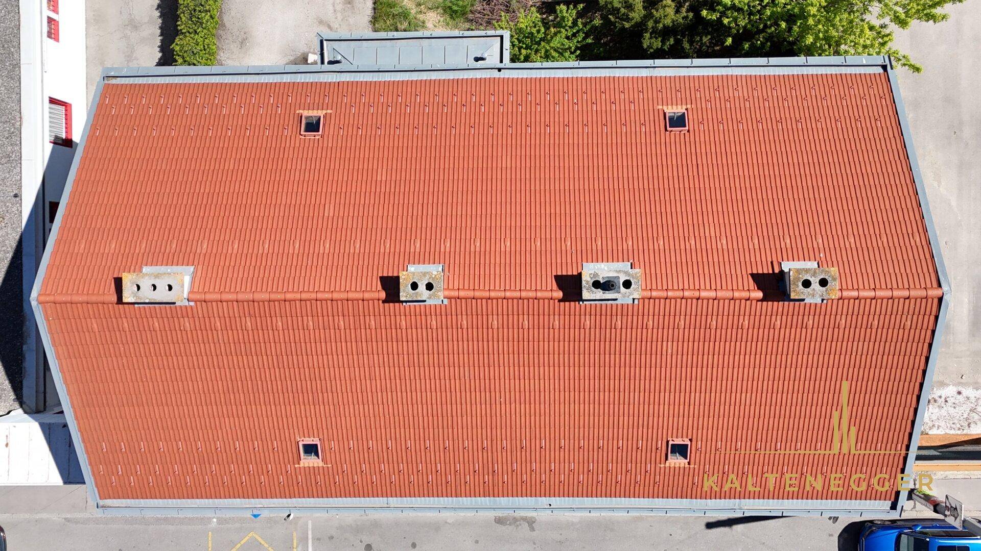 Draufsicht auf ein Gebäude mit rotem Ziegeldach und mehreren Dachfenstern bei klarem Himmel.