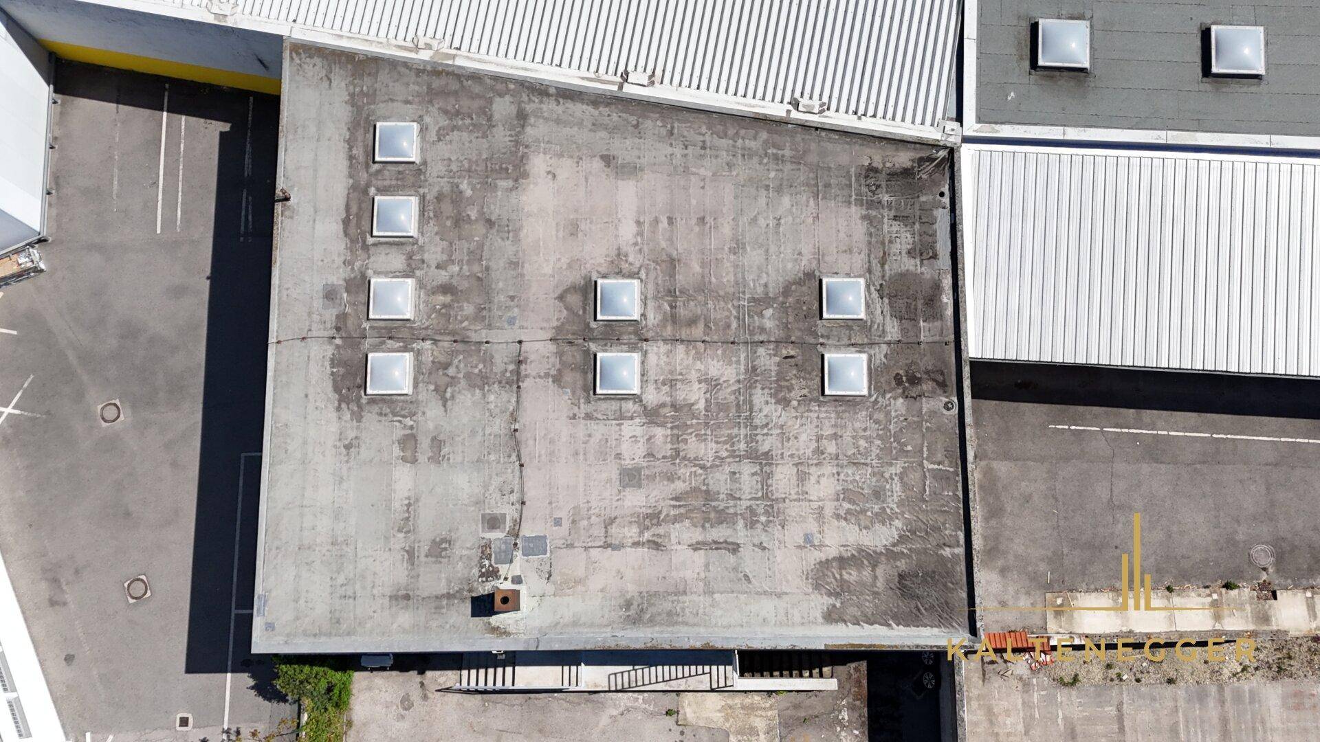 Draufsicht auf ein Flachdach mit mehreren Oberlichtern und angrenzenden Gebäudeteilen.