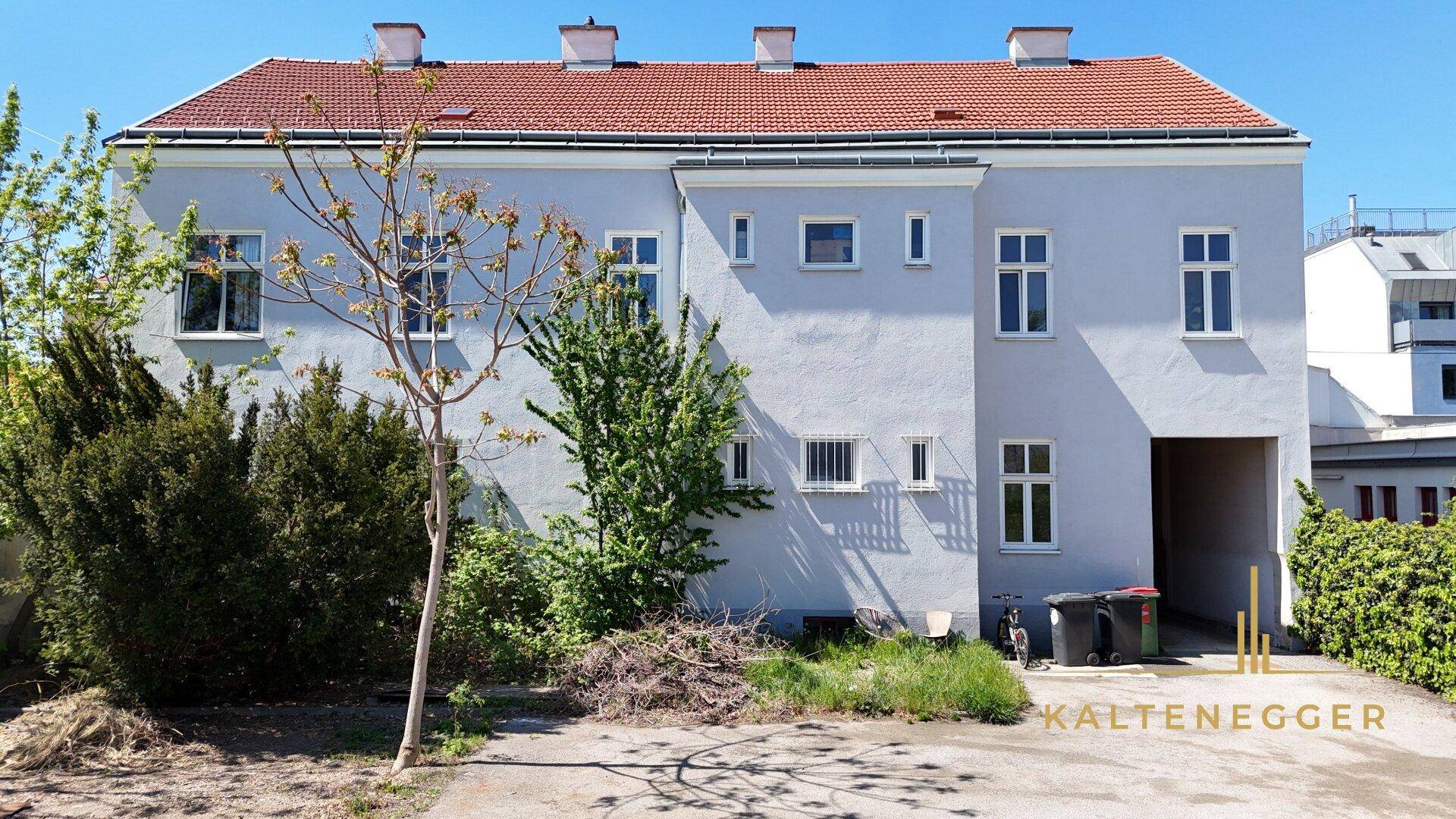 Rückansicht eines Gebäudes mit hellgrauer Fassade, roten Dachziegeln und ungepflegtem Gartenbereich.