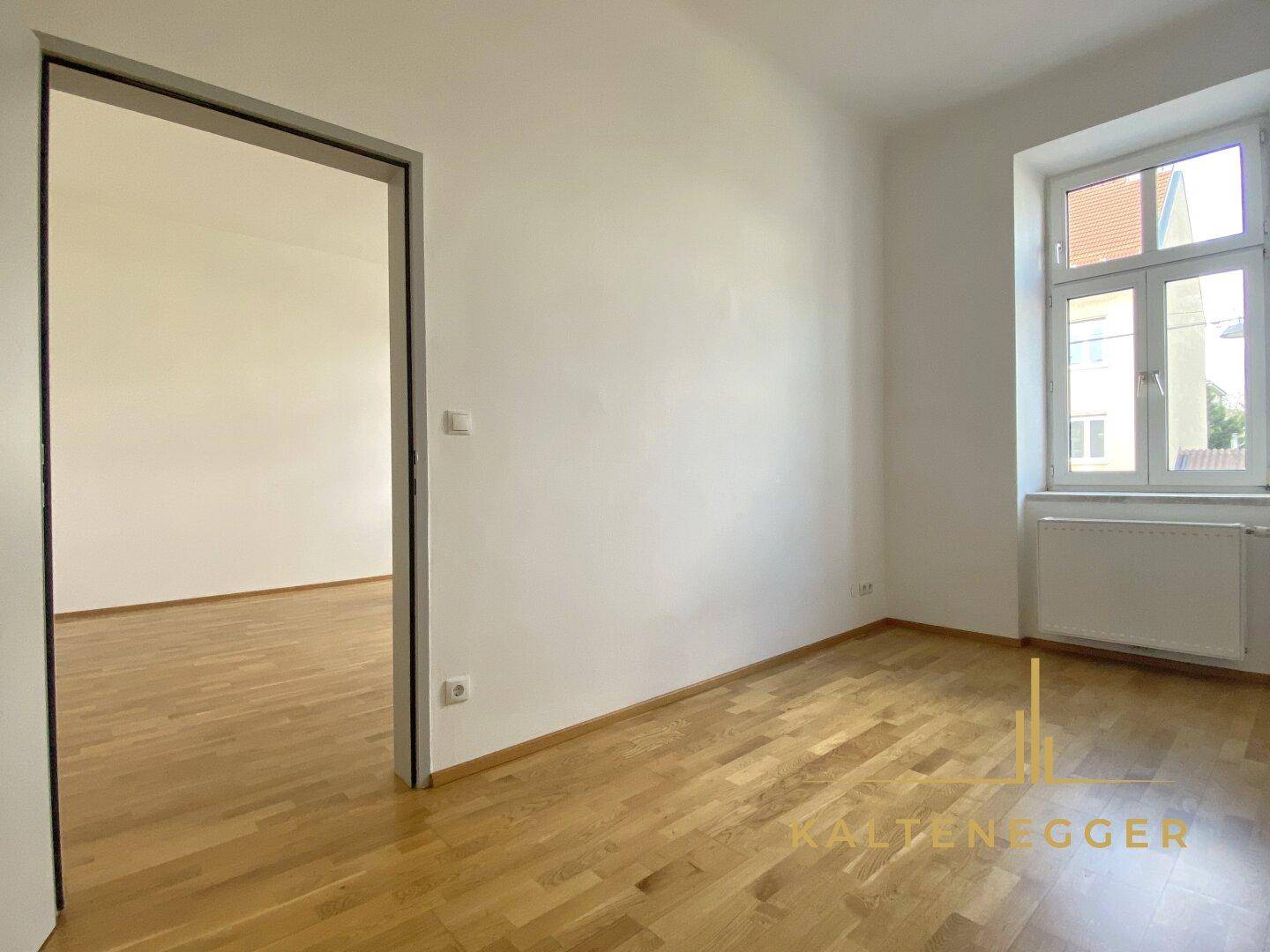Leeres Schlafzimmer mit Parkettboden und einem Fenster, viel Tageslicht.