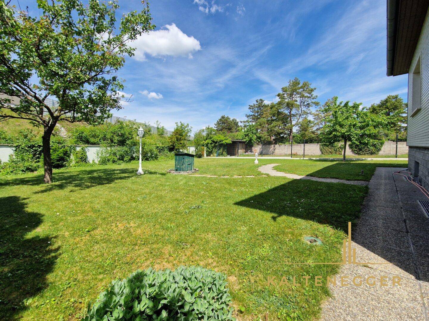 Weitläufiger Garten mit Rasenfläche, Bäumen und einem geschwungenen Weg unter blauem Himmel.