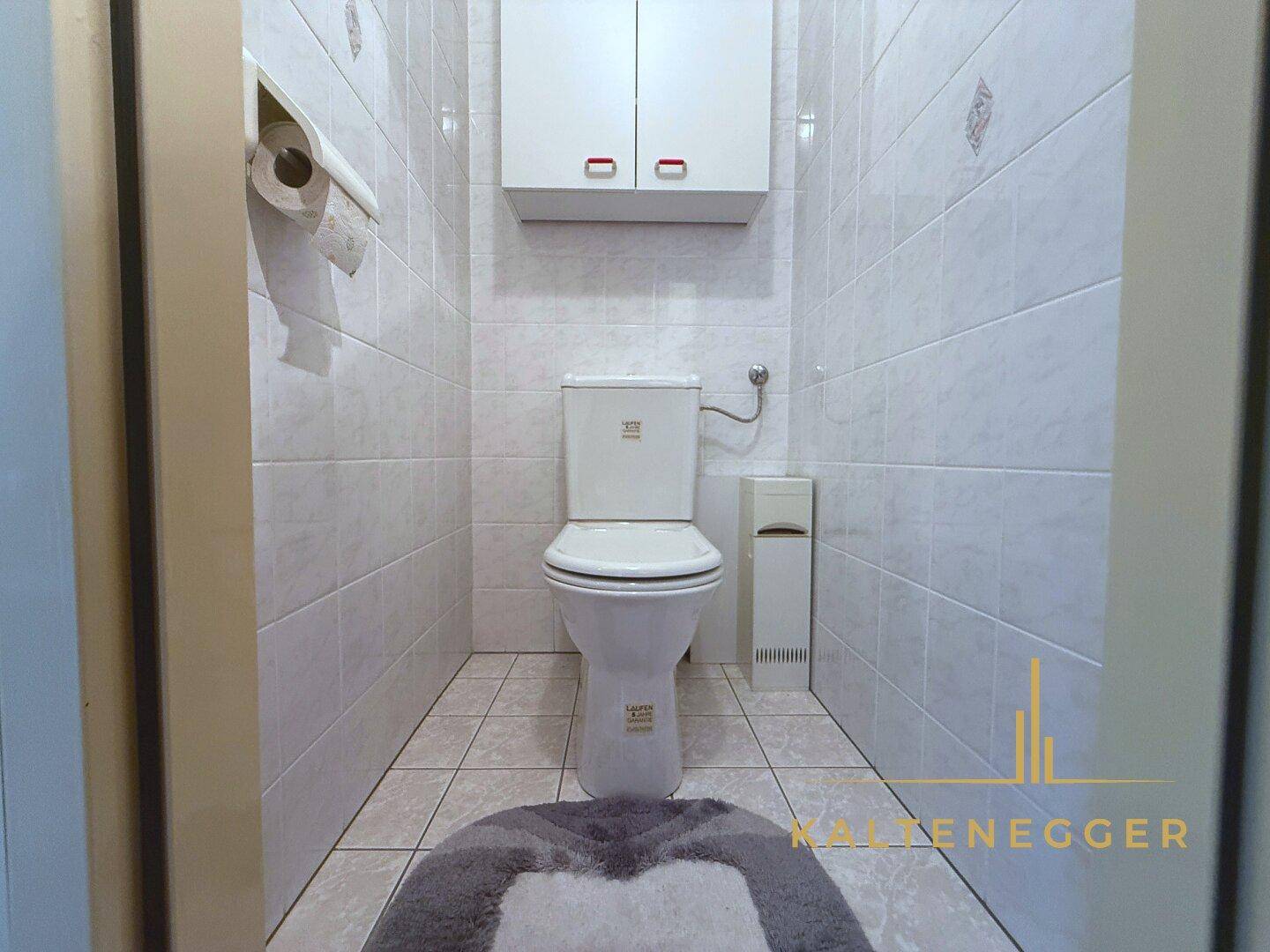 Separates WC mit Toilette und kleinem Hängeschrank, vollständig gefliest.
