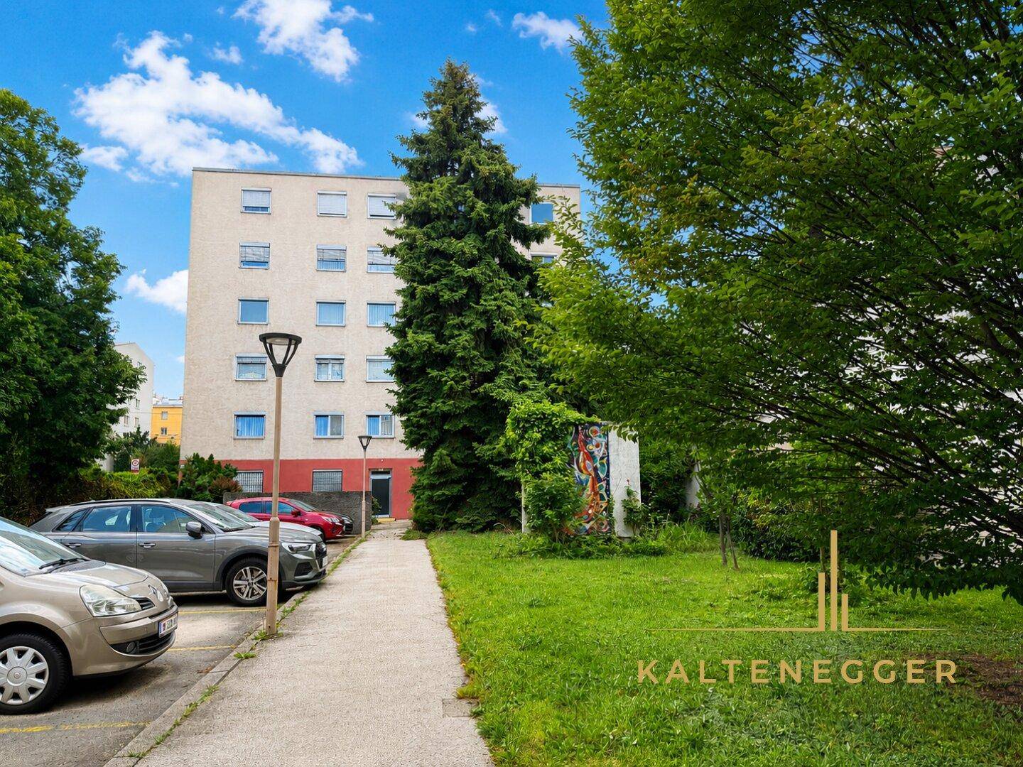 Mehrfamilienhaus in grüner Umgebung mit Parkplätzen und hohen Bäumen.