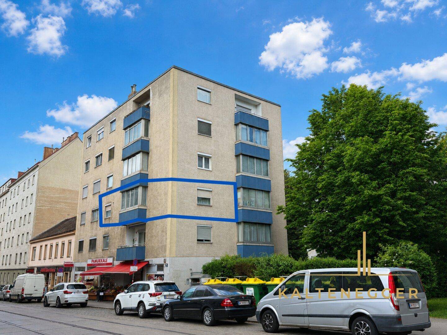 Ansicht des Mehrfamilienhauses von der Straße mit Balkonen und Geschäften im Erdgeschoss.