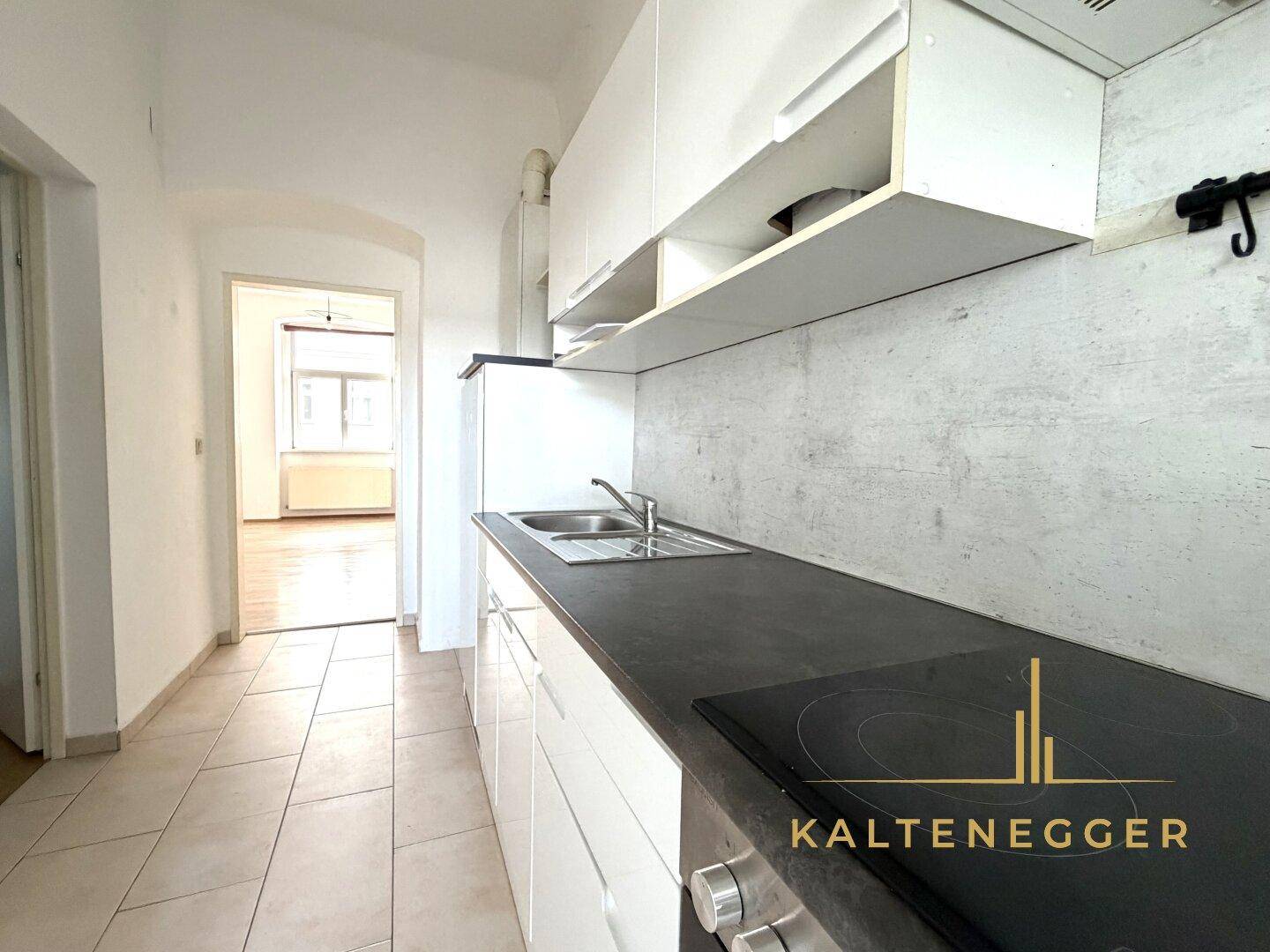 Kleine Küche mit weißen Schränken, dunkler Arbeitsplatte und Fliesenboden, mit Blick in einen weiteren Raum.