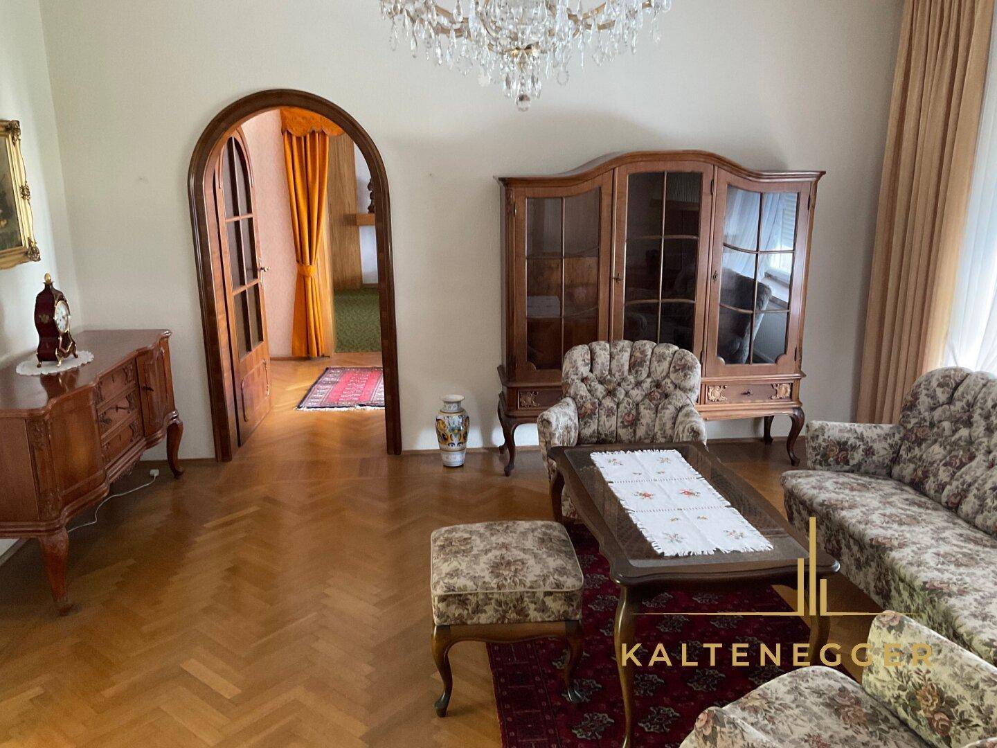 Gemütliches Wohnzimmer mit klassischem Mobiliar, Parkettboden und Zugang zu einem weiteren Raum.