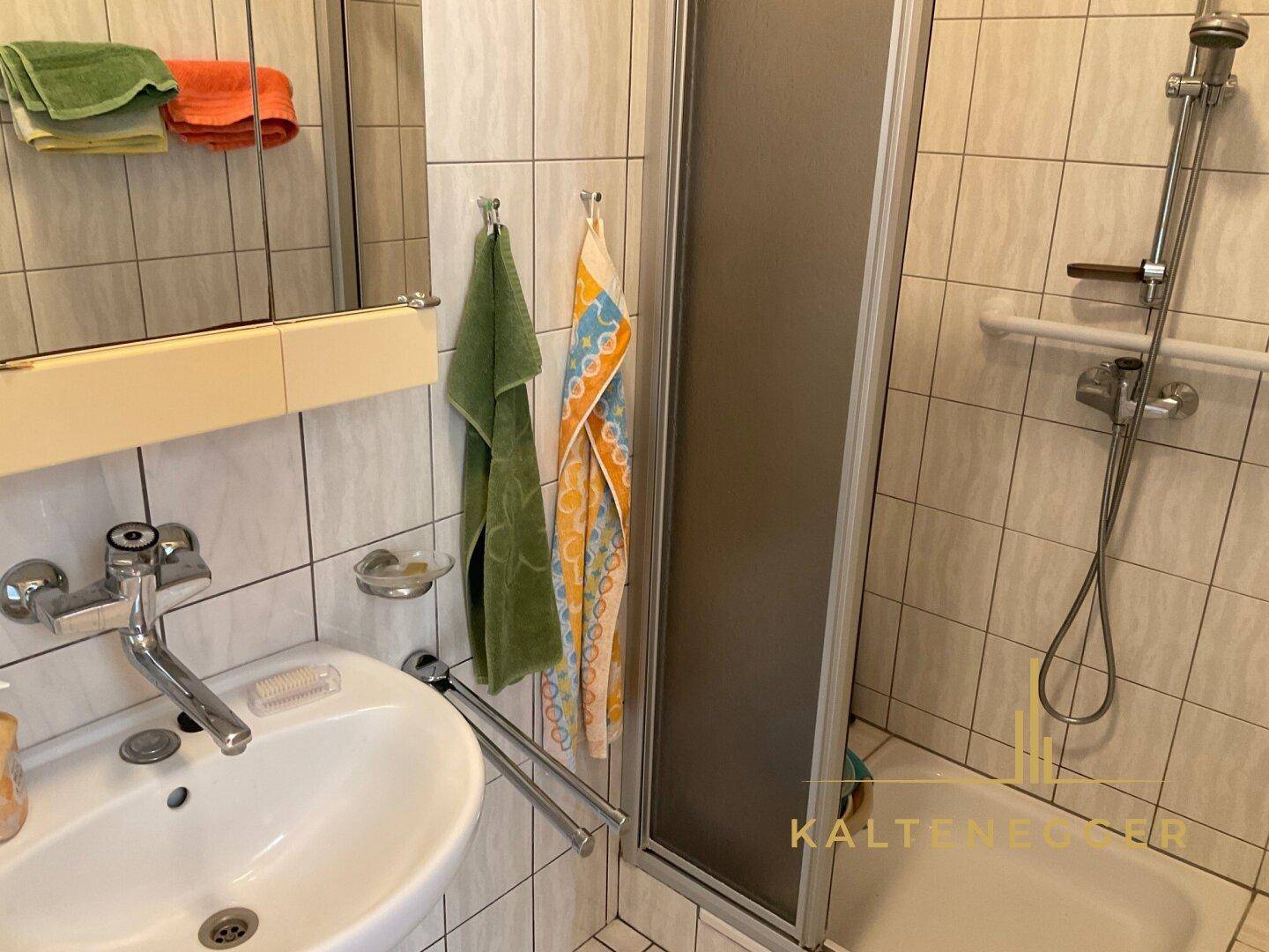 Kleines Badezimmer mit Dusche, Waschbecken und hellen Fliesen, ausgestattet mit Haltegriffen.