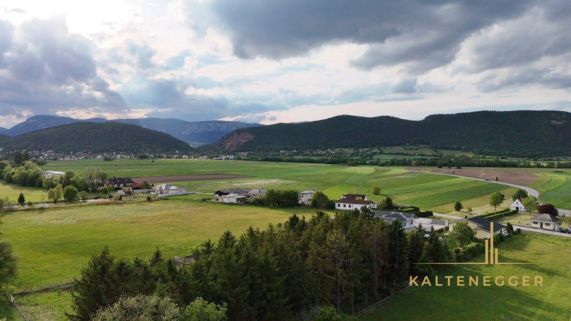 Luftaufnahme eines Tals mit grünen Feldern, Häusern und bewaldeten Bergen im Hintergrund unter bewölktem Himmel.