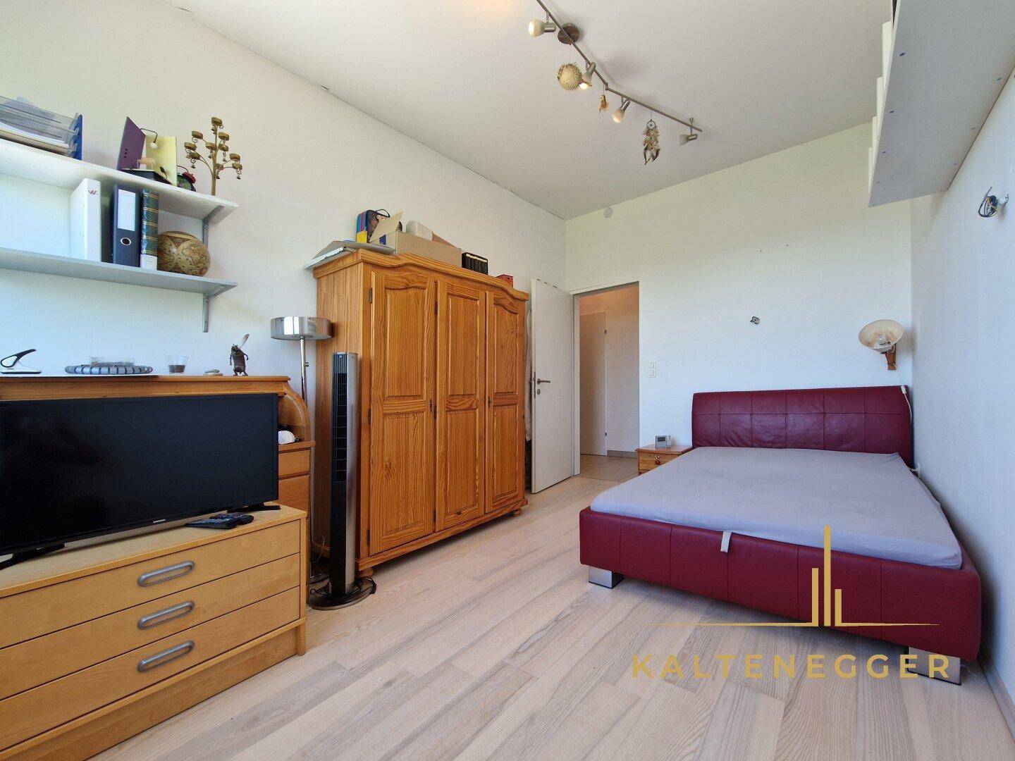 Gemütliches Schlafzimmer mit Holzboden, Holzschrank und rotem Bett, gut beleuchtet.