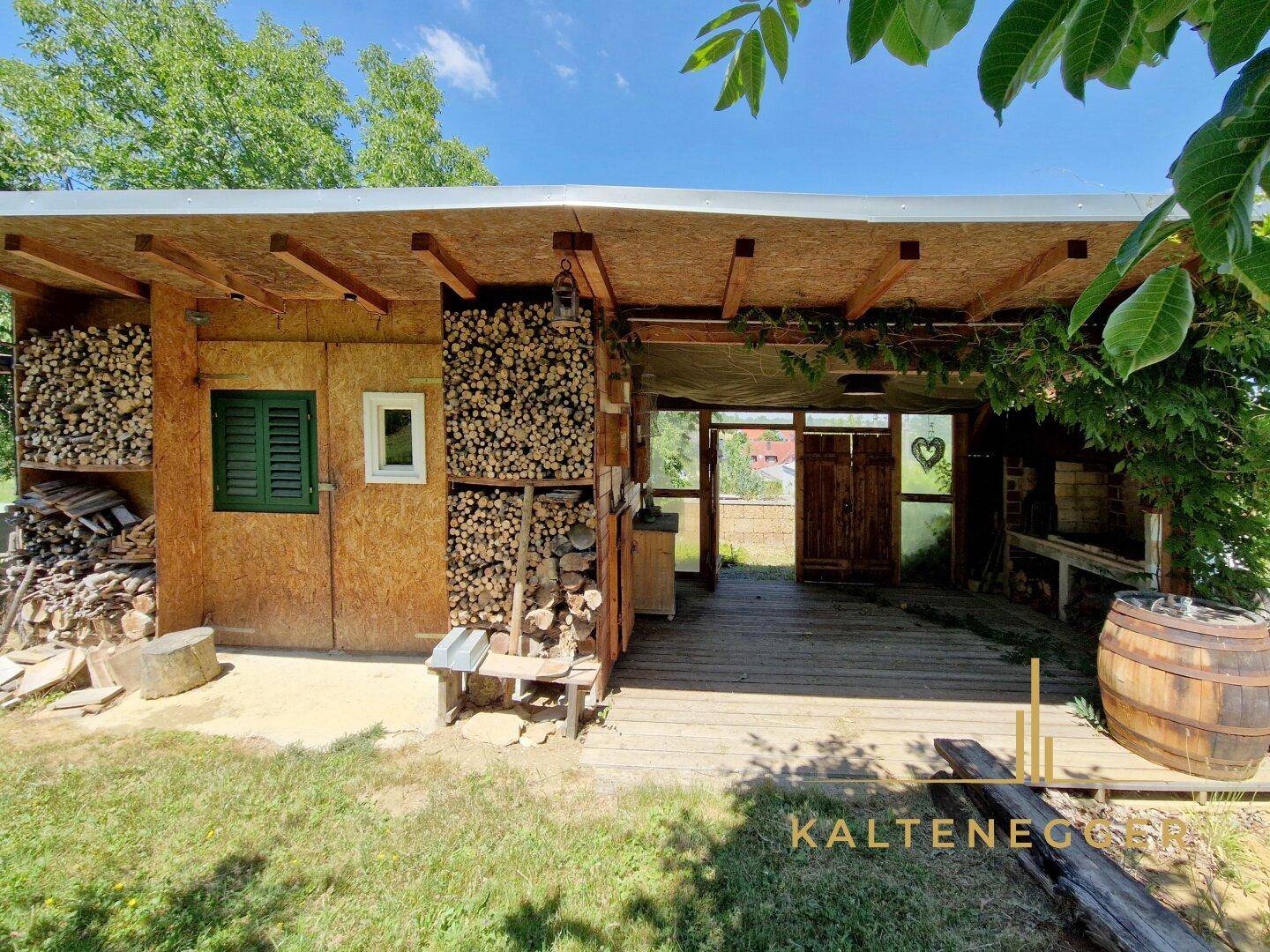 Rustikaler überdachter Außenbereich mit Holzstapeln und einer kleinen Terrasse, umgeben von Grün.