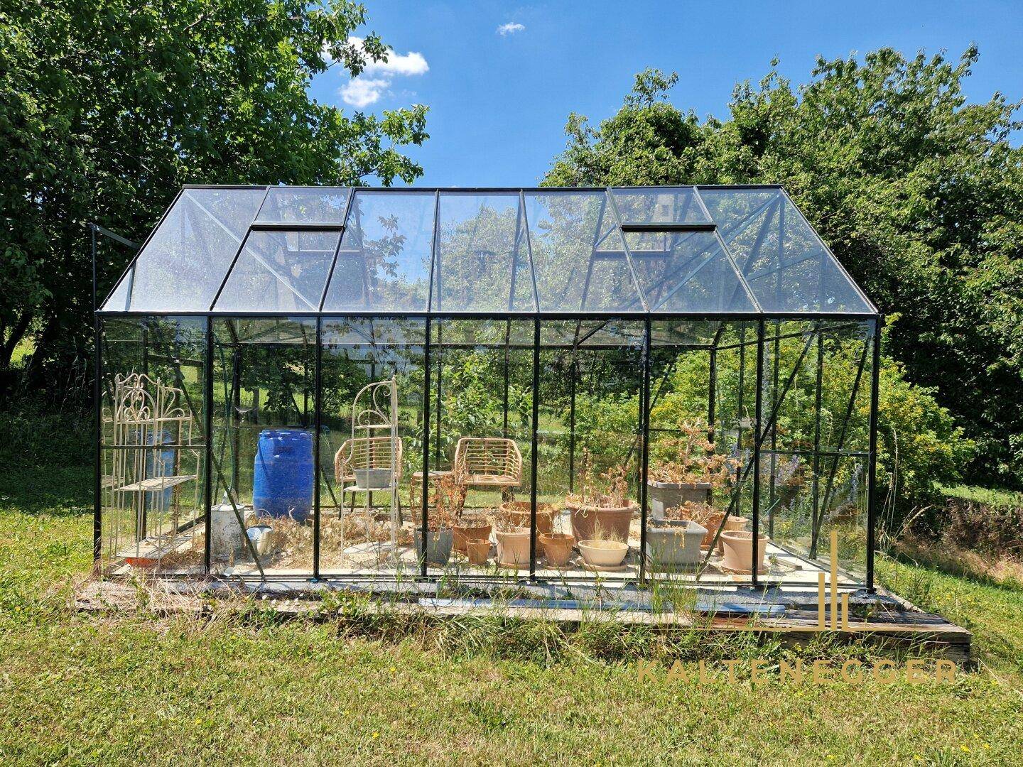 Modernes Gewächshaus aus Glas und Metall, gefüllt mit Pflanzen und Gartenutensilien, auf einer Rasenfläche.