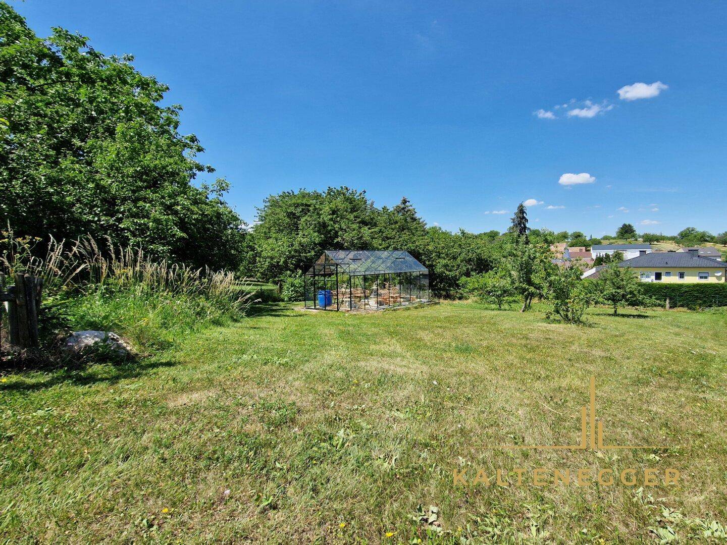 Weitläufiger Garten mit großer Rasenfläche und einem Gewächshaus im Hintergrund unter blauem Himmel.