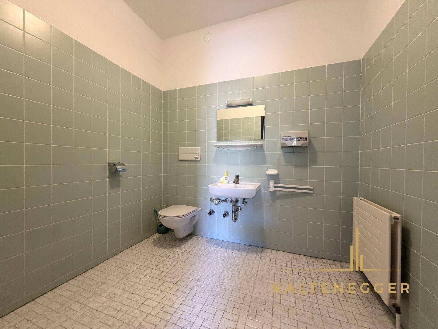 Badezimmer mit hellgrünen Wandfliesen, weißem Fliesenboden und barrierefreien Haltegriffen.