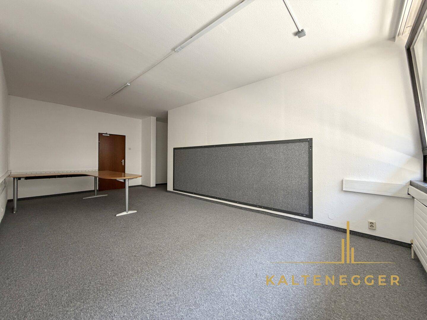 Büro mit dunklem Teppichboden, einer großen Pinnwand und einem Arbeitstisch.