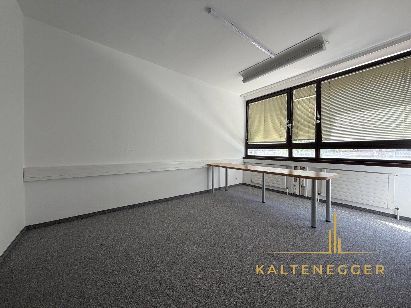 Helles Büro mit grauem Teppichboden, langen Tischen und Fensterjalousien für Privatsphäre.