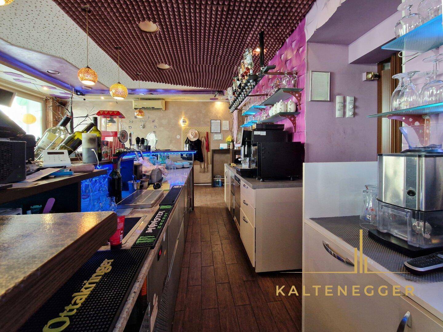 Blick hinter die Bar mit professionellen Geräten, Flaschenregalen und Arbeitsflächen für den Service.