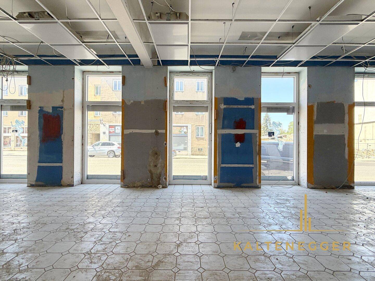 Ladenlokal mit großen Schaufenstern, Fliesenboden und sichtbaren Renovierungsspuren an den Wänden.