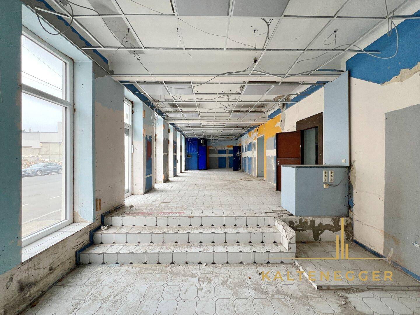 Langer Verkaufsraum mit Stufen, Fliesenboden und einer offenen Deckenkonstruktion, die renoviert werden muss.