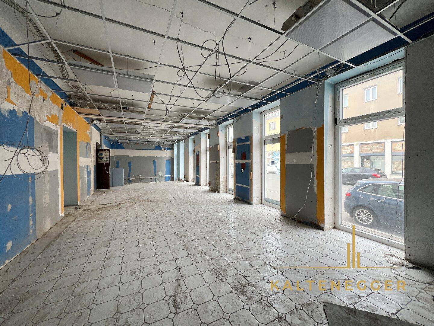 Weitläufiger Gewerberaum mit Fliesenboden, großen Fenstern und sichtbaren Deckeninstallationen, die erneuert werden müssen.
