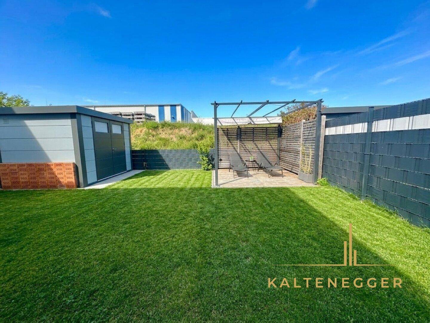 Gepflegter Gartenbereich mit Rasenfläche und einer kleinen Terrasse mit Liegestühlen, umgeben von einem Zaun.