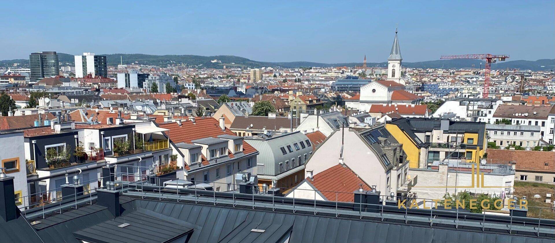 Weitläufiger Stadtblick über Dächer und Gebäude mit Kirchturm und Bergen im Hintergrund.