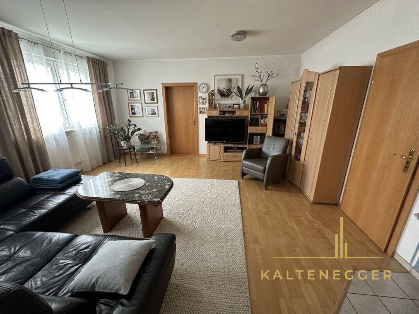 Geräumiges Wohnzimmer mit schwarzem Ledersofa, hellem Parkettboden und einem TV-Möbel.